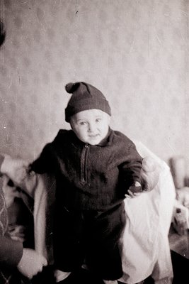 Vintage black-and-white portrait of a toddler in a knitted hat and long-sleeve sweater, seated indoors. The child’s expressio...