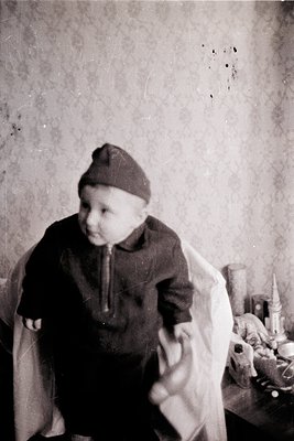 Vintage sepia-toned portrait of a toddler in a knitted cap and long-sleeve shirt, seated indoors near a table with jars and a...