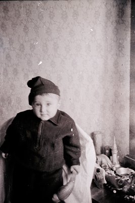 Vintage black-and-white portrait of a young child in early 20th-century attire—knitted cap, long-sleeved sweater, and a drape...