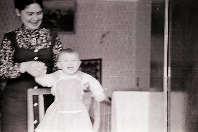 A mid-20th century black-and-white photo of a woman guiding a toddler in a highchair indoors, likely a home setting. The woma...