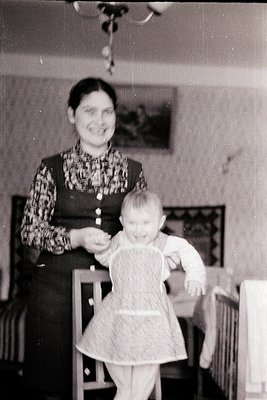 Mid-20th century indoor portrait: woman in patterned blouse and dark vest guides a toddler in a white dress, seated on a high...