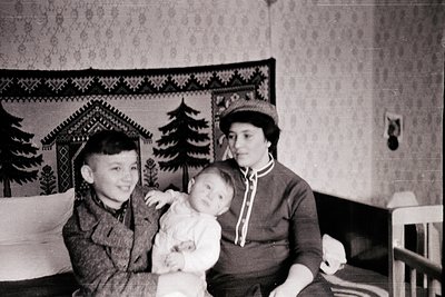Vintage black-and-white portrait of a woman holding a baby, with a young boy seated beside her in a modest indoor setting. De...