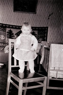 Toddler in vintage dress stands on wooden chair in mid-20th-century interior, holding a small object. Wallpaper features flor...