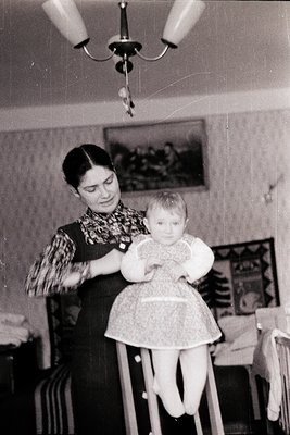 Vintage black-and-white photo of a woman in a patterned blouse and dark skirt holding a toddler in a highchair, likely mid-20...