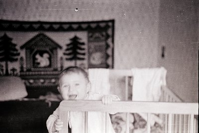 Vintage black-and-white photo of a toddler leaning on a wooden crib rail, smiling directly at the camera. Decorative wallpape...