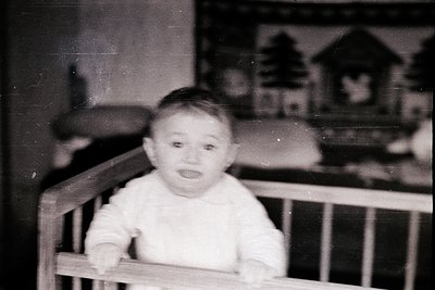 Vintage black-and-white photo of a toddler in a wooden crib, wearing a long-sleeve shirt. Decorative wallpaper with Asian-ins...