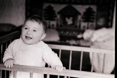 Mid-20th century black-and-white photo of a toddler in a crib, smiling at the camera. The child wears a short-sleeved, high-n...