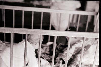Vintage black-and-white shot of a child’s crib with vertical metal bars, blurred face partially visible. Soft fabric and bedd...