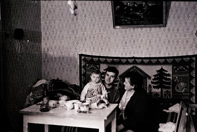 Vintage black-and-white interior shot of a family—man, woman, and child—seated at a simple wooden table in a modestly decorat...