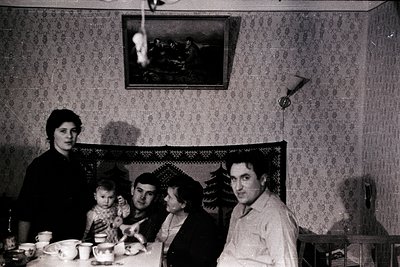 Family portrait in a modestly decorated 1960s–70s living room, featuring patterned wallpaper and a framed landscape painting....
