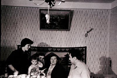 Vintage black-and-white interior shot of a mid-20th-century gathering. Five people seated around a table in a dimly lit room ...