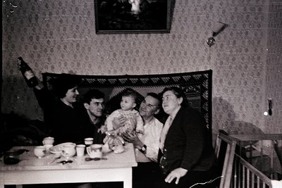 Vintage black-and-white indoor family portrait, likely late 20th century. Three adults and a baby pose around a small table w...