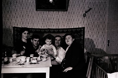 Family portrait from mid-20th century, likely 1950s–1960s. Five individuals seated at a table in a modest indoor setting, fra...
