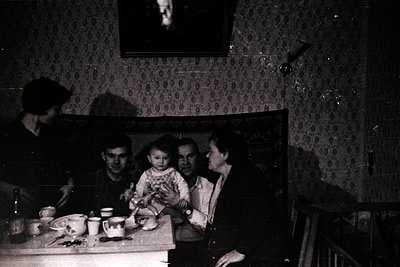 Family portrait in dimly lit room, likely mid-20th century. Four adults and a child seated around a small wooden table with b...