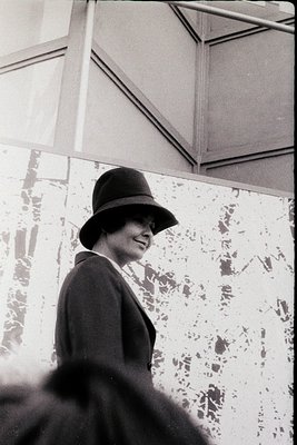 Black-and-white portrait of a woman in a fedora and dark coat, smiling at the camera. Architectural details include a ceiling...