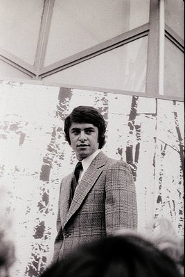 Young man in a 1970s-style checkered blazer and dress shirt poses indoors, likely at a formal event or gathering. The pattern...