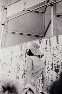 Vintage black-and-white shot of a woman in a wide-brimmed hat and striped blouse walking under a modernist overpass, likely m...