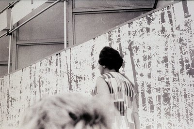 Black-and-white photo of a woman in a striped dress examining a large mural or wall art covered in abstract, textured pattern...