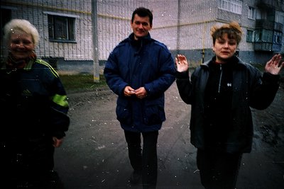 Three individuals pose outdoors in light rain, framed by a blurred, monochromatic urban backdrop. The man in the center wears...