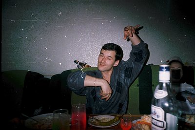 Vintage indoor scene featuring a man in 1970s-style clothing raising a glass in a dimly lit room. Patterned wallpaper and a g...