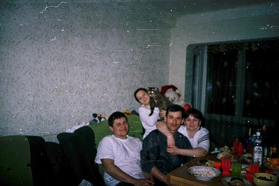 Vintage indoor family portrait in a modestly decorated living room, likely 1980s–1990s. Four people pose on a green sofa agai...