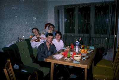 Family gathering at a wooden dining table in a dimly lit room, likely 1980s–1990s Eastern Europe. Four adults pose with food,...