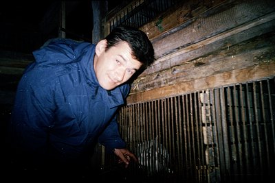 Man in a dark blue raincoat leans against a weathered wooden fence under low-light conditions, possibly nighttime. The fence’...