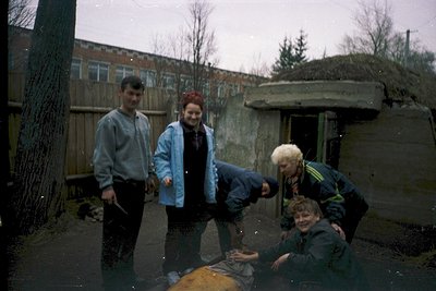 Four individuals pose outdoors in wet conditions, likely post-rain, during the 1980s–1990s. The man on the left wears a light...