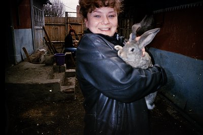 A woman in a dark raincoat holds a young rabbit indoors near a rustic farm setting. Wooden structures, metal feeders, and a w...