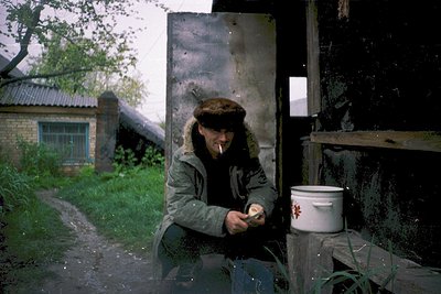 Vintage photograph of a man in a rural setting, repairing a rusted metal door with a wrench. He wears a knitted cap and heavy...