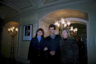 Three adults pose indoors in a grand, dimly lit hall with ornate chandeliers and classical architecture. The man wears a dark...
