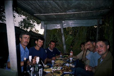 Vintage group photo under a makeshift canopy, likely late 20th century. Five men seated around a table laden with food (meat,...