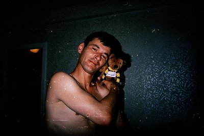 Vintage-style portrait of a shirtless man holding a vintage teddy bear in a dimly lit bathroom. Patterned wallpaper with flor...