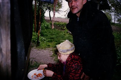 Vintage photo of two individuals in a rural setting, likely Eastern Europe. A man in a dark jacket leans over a wooden struct...