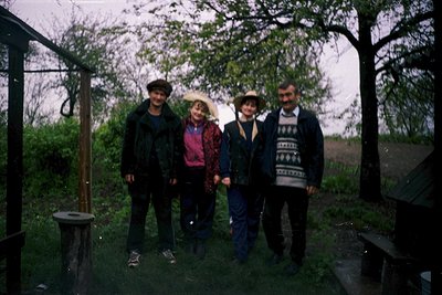 Vintage group portrait in rural outdoor setting, likely late 20th century. Four individuals pose under a wooden pergola surro...