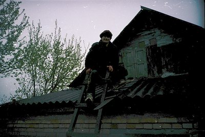 Vintage black-and-white photo of a person in winter attire climbing a ladder on a rustic wooden house with a corrugated metal...