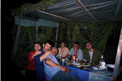 Group of six seated under a canopy tent at night, likely enjoying a meal outdoors. Table set with plates, drinks, and a bottl...