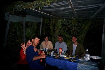 Vintage group photo under a rustic metal pavilion draped in greenery. Five adults seated at a blue table, holding drinks and ...