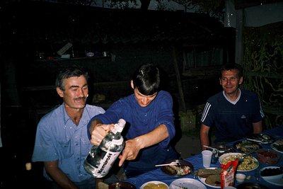 Vintage indoor gathering featuring three men in 1970s-style attire—light blue button-ups and Adidas tracksuits—around a table...