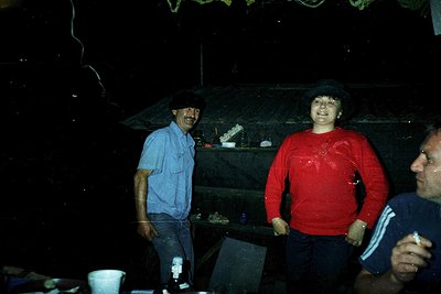 Vintage indoor photo of three individuals in casual 1970s attire—man in light blue shirt, woman in red sweater, and partial v...