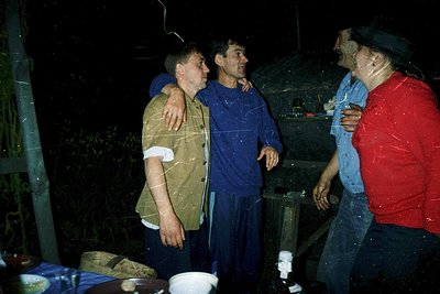 Three men in wet, casual attire share a moment indoors during heavy rain, likely in a rural setting. The man in blue stands c...