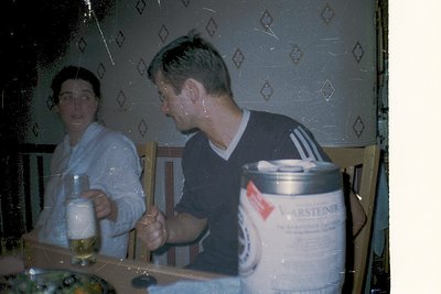Vintage indoor scene featuring two people seated at a wooden table. The man wears a navy Adidas sweatshirt and holds a glass,...
