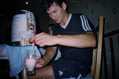 Vintage indoor shot of a man in a striped Adidas shirt pouring beer from a bottle into a glass, using a small funnel. A large...