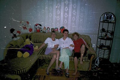 Vintage indoor family portrait from the 1970s–1980s, featuring three adults seated on a patterned sofa. Decor includes Soviet...