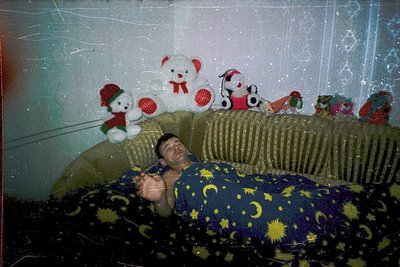 Vintage photo of a man reclining on a bed adorned with retro celestial-patterned bedding. Surrounding him are plush toys—incl...