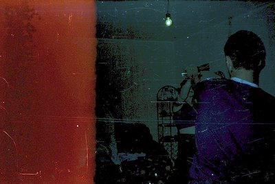 Vintage film grain overlay reveals a dimly lit interior with a man in a dark suit facing away, holding a cigarette. Faded red...