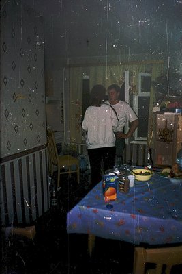 Two individuals stand in a dimly lit kitchen, reflected in a foggy mirror. The man wears a white shirt and dark pants; the wo...
