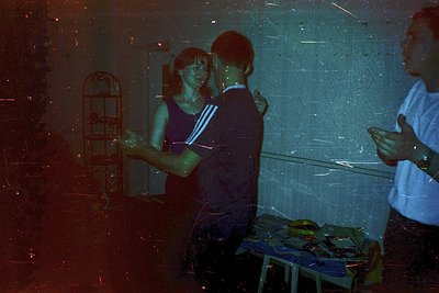 Vintage indoor scene with three individuals in a dimly lit room, likely a 1970s-1980s setting. A woman in a striped tank top ...