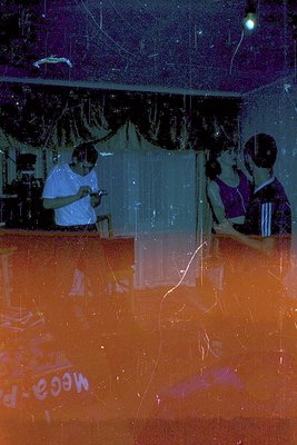 Reflection of a dimly lit indoor party with orange-tinted floor. Two blurred figures—one in white (left) and two in purple/wh...