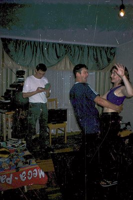 Vintage indoor party scene with confetti-covered guests. A man in a white shirt holds a camera, while a woman in a purple tan...